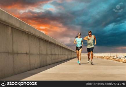 fitness, sport and healthy lifestyle concept - happy couple in sports clothes and sunglasses running along pier over sunset sky background. couple in sports clothes running along pier