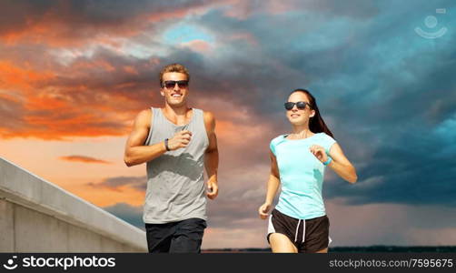 fitness, sport and healthy lifestyle concept - happy couple in sports clothes and sunglasses running over sunset sky background. couple in sports clothes running outdoors