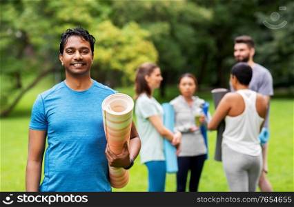 fitness, sport and healthy lifestyle concept - happy smiling indian young man with mat over group of people meeting for yoga class at summer park. smiling man with yoga mat over group of people