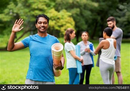 fitness, sport and healthy lifestyle concept - happy smiling indian young man with mat waving hand over group of people meeting for yoga class at summer park. smiling man with yoga mat over group of people