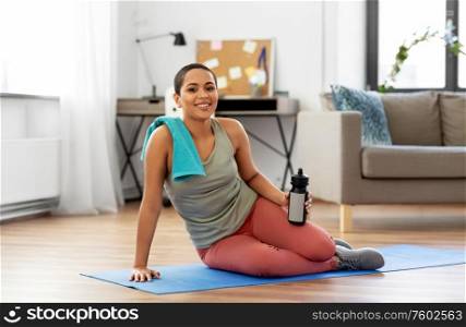 fitness, sport and healthy lifestyle concept - happy smiling young african american woman with bottle of water and towel stting on mat at home. woman with bottle and towel after sports at home