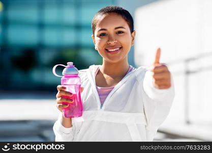 fitness, sport and healthy lifestyle concept - happy smiling young african american woman with bottle of water showing thumbs up outdoors. happy woman with bottle of water showing thumbs up