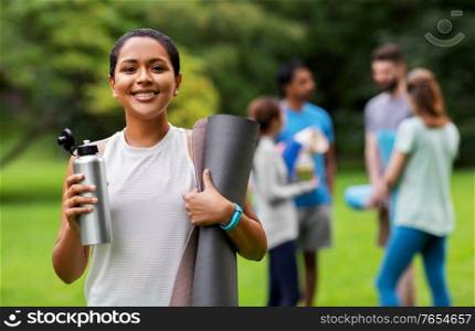 fitness, sport and healthy lifestyle concept - happy smiling young african american woman with mat and bottle over group of people meeting for yoga class at summer park. smiling woman with yoga mat and bottle at park