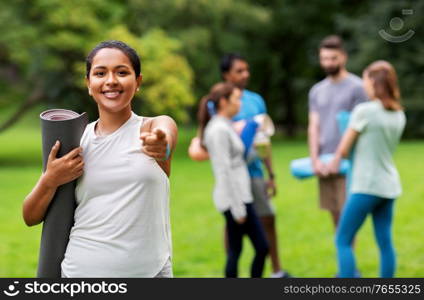 fitness, sport and healthy lifestyle concept - happy smiling young african american woman with mat pointing finger to camera over group of people meeting for yoga class at summer park. woman with yoga mat pointing finger to camera