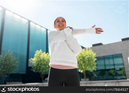 fitness, sport and healthy lifestyle concept - happy smiling young african american woman stretching arm outdoors. african american woman doing sports outdoors