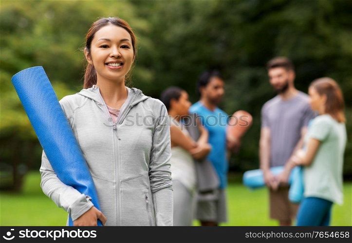 fitness, sport and healthy lifestyle concept - happy smiling young asian woman with mat over group of people meeting for yoga class at summer park. smiling woman with yoga mat over group of people