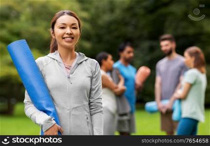 fitness, sport and healthy lifestyle concept - happy smiling young asian woman with mat over group of people meeting for yoga class at summer park. smiling woman with yoga mat over group of people