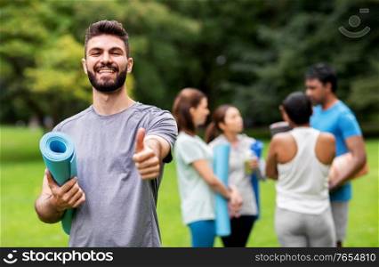 fitness, sport and healthy lifestyle concept - happy smiling young man with mat showing thumbs up over group of people meeting for yoga class at summer park. smiling man with yoga mat showing thumbs up