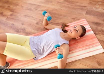 fitness, sport and healthy lifestyle concept - happy teenage girl with dumbbells exercising on yoga mat at home. teenage girl with dumbbells exercising at home