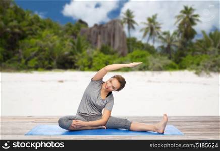 fitness, sport and healthy lifestyle concept - happy woman doing yoga and stretching on mat over exotic tropical beach background. happy woman doing yoga and stretching on beach