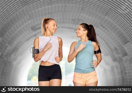 fitness, sport and healthy lifestyle concept - happy young women or female friends with earphones wearing armbands with smartphones and running outdoors. young women with earphones and smartphones running