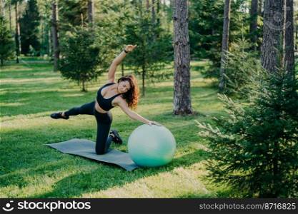 Fitness, sport and healthy lifestyle concept. Outdoor shot of slim brunette European woman does pilates and balance exercise with fit ball tilts aside poses in green forest on grass at karemat.