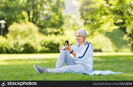 fitness, sport and healthy lifestyle concept - senior woman with earphones listening to music on smartphone sitting on exercise mat at summer park. sporty senior woman with earphones and smartphone