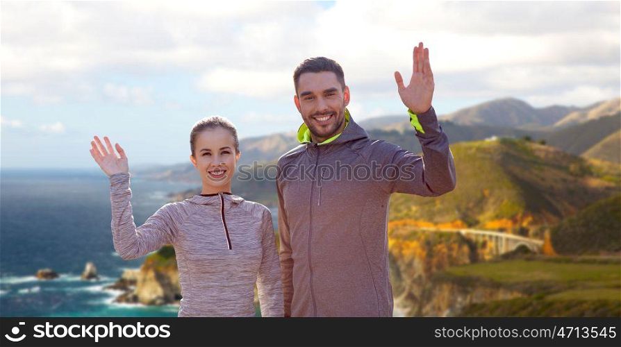 fitness, sport, and healthy lifestyle concept - smiling couple waving hand over bixby creek bridge on big sur coast of california background. smiling couple in sport clothes waving hand. smiling couple in sport clothes waving hand