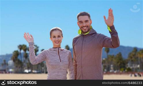 fitness, sport, and healthy lifestyle concept - smiling couple waving hand over venice beach background in california. smiling couple in sport clothes waving hand. smiling couple in sport clothes waving hand