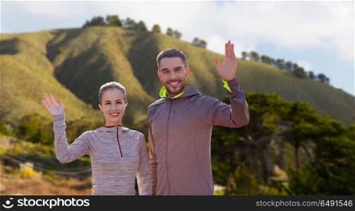 fitness, sport, and healthy lifestyle concept - smiling couple waving hand over big sur hills background in california. smiling couple in sport clothes waving hand. smiling couple in sport clothes waving hand