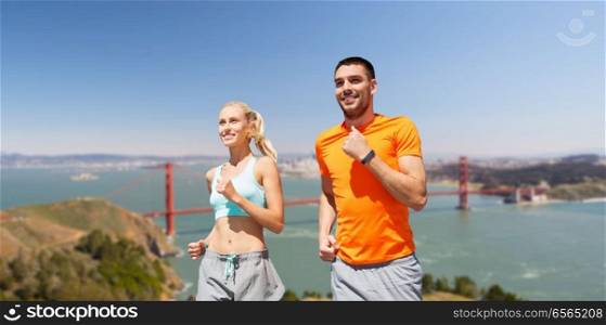 fitness, sport and healthy lifestyle concept - smiling couple with heart-rate watch running over golden gate bridge in san francisco bay background. happy couple running over golden gate bridge