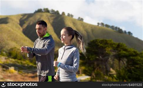 fitness, sport and healthy lifestyle concept - smiling couple with heart-rate watches running over big sur hills background in california. couple with fitness trackers running outdoors. couple with fitness trackers running outdoors