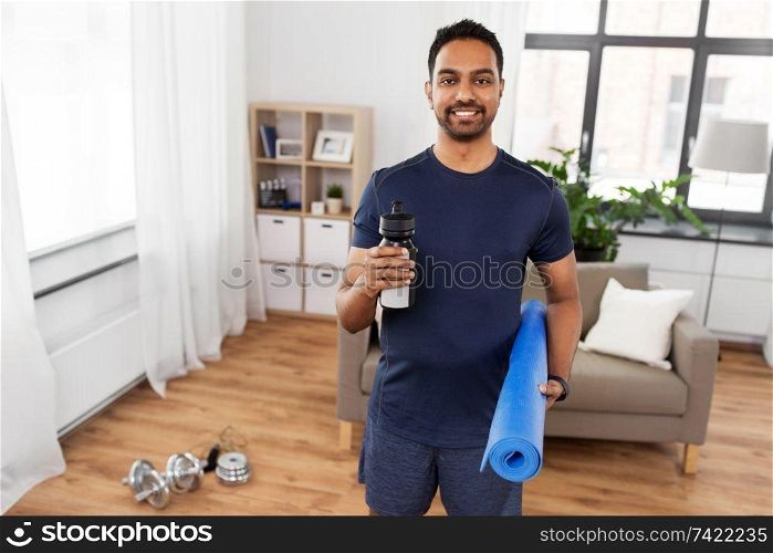 fitness, sport and healthy lifestyle concept - smiling indian man with exercise mat and bottle of water or energy drink at home. indian man with exercise mat and bottle at home