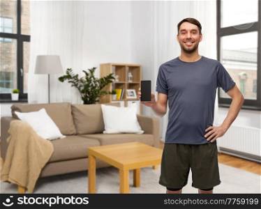 fitness, sport and healthy lifestyle concept - smiling man in sports clothes showing smartphone over home room background. smiling man in sports clothes showing smartphone