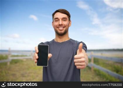 fitness, sport and healthy lifestyle concept - smiling man in sports clothes showing smartphone and thumbs up gesture over natural background. smiling man in sports clothes showing smartphone