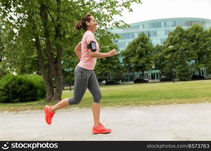 fitness, sport and healthy lifestyle concept - smiling woman with earphones wearing armband with smartphone and jogging at park and listening to music. woman with earphones add armband jogging at park