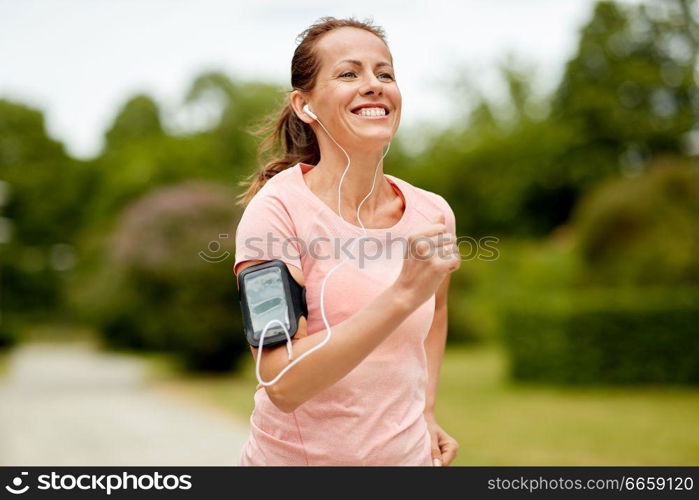 fitness, sport and healthy lifestyle concept - smiling woman with earphones wearing armband for smartphone, jogging at summer park and listening to music. woman with earphones add armband jogging at park