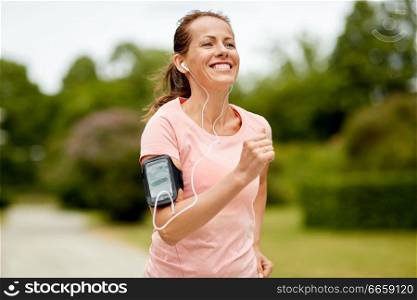 fitness, sport and healthy lifestyle concept - smiling woman with earphones wearing armband for smartphone, jogging at summer park and listening to music. woman with earphones add armband jogging at park