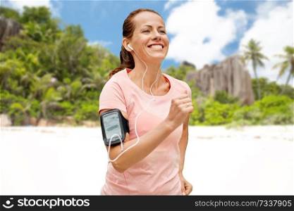 fitness, sport and healthy lifestyle concept - smiling woman with earphones wearing armband for smartphone, running and listening to music over tropical beach on seychelles island background. woman with earphones and armband running on beach