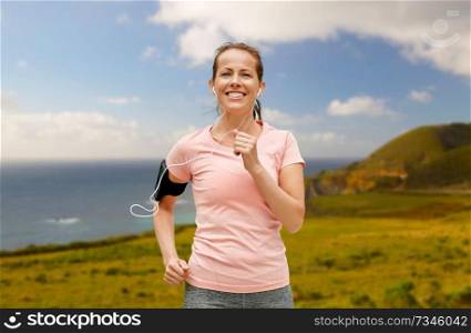 fitness, sport and healthy lifestyle concept - smiling woman with earphones wearing armband for smartphone, running and listening to music over big sur coast of california background. woman with earphones and armband running outdoors