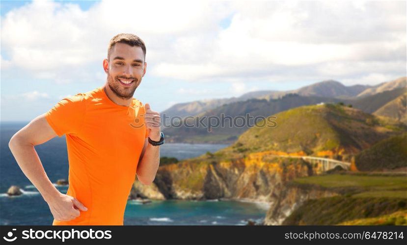fitness, sport and healthy lifestyle concept - smiling young man with heart rate watch running over big sur hills and pacific ocean background in california. smiling young man running at summer seaside. smiling young man running at summer seaside