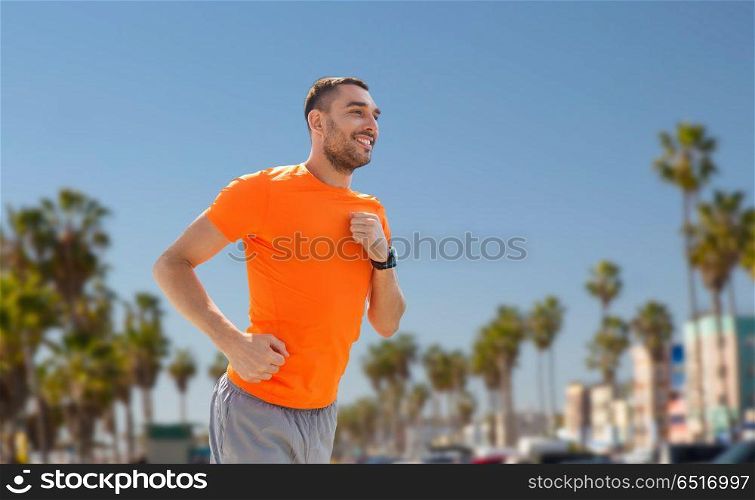 fitness, sport and healthy lifestyle concept - smiling young man with heart rate watch running over venice beach background in california. smiling young man running at summer seaside. smiling young man running at summer seaside