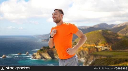 fitness, sport and healthy lifestyle concept - smiling young man with heart rate watch running over big sur hills and pacific ocean background in california. smiling young man running at summer seaside. smiling young man running at summer seaside