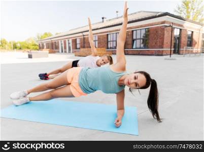 fitness, sport and healthy lifestyle concept - smiling young women or female friends doing side plank with raised hand outdoors together. women doing side plank on sport mats outdoors
