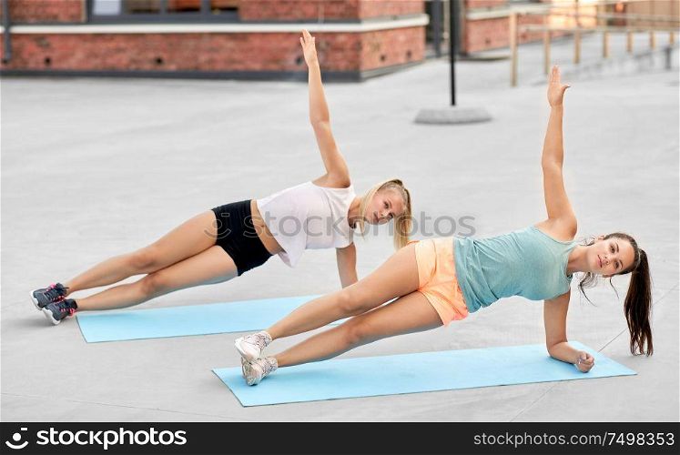 fitness, sport and healthy lifestyle concept - smiling young women or female friends doing side plank outdoors together. women doing side plank on sport mats outdoors