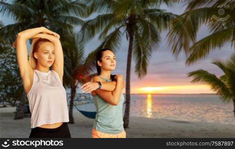 fitness, sport and healthy lifestyle concept - smiling young women or female friends with activity trackers stretching over tropical beach on background. women with fitness trackers stretching on beach