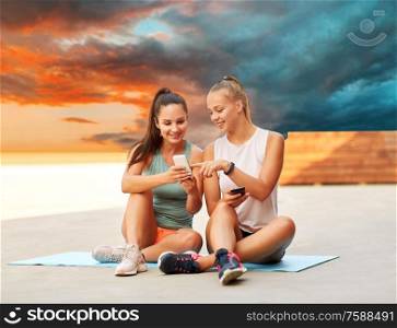 fitness, sport and healthy lifestyle concept - smiling young women or female friends with smartphones on roof top over sunset sky on background. sporty women or friends with smartphone