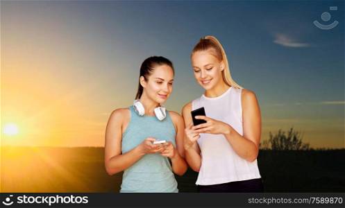 fitness, sport and healthy lifestyle concept - smiling young women or female friends with smartphones over sunset sky on background. women or female friends with smartphones