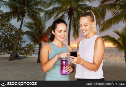 fitness, sport and healthy lifestyle concept - smiling young women or female friends with smartphone and flask over palm trees on tropical beach on background. women or female friends with smartphone and flask