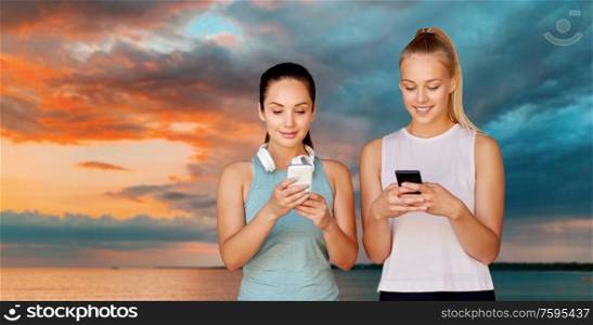 fitness, sport and healthy lifestyle concept - smiling young women or female friends with smartphones over sea and sunset sky on background. women or female friends with smartphones