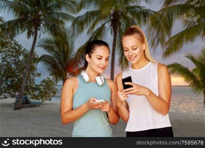 fitness, sport and healthy lifestyle concept - smiling young women or female friends with smartphones over tropical beach on background. women or female friends with smartphones on beach