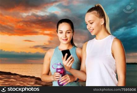 fitness, sport and healthy lifestyle concept - smiling young women or female friends with smartphone and flask over sea and sunset sky on background. women or female friends with smartphone and flask