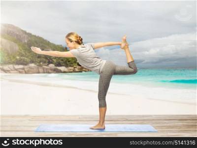 fitness, sport and healthy lifestyle concept - woman doing yoga in lord of the dance pose on mat over exotic tropical beach background. woman doing yoga lord of the dance pose on beach