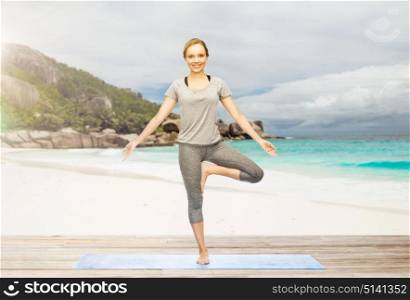 fitness, sport and healthy lifestyle concept - woman doing yoga in tree pose on mat over exotic tropical beach background. woman doing yoga in tree pose on beach