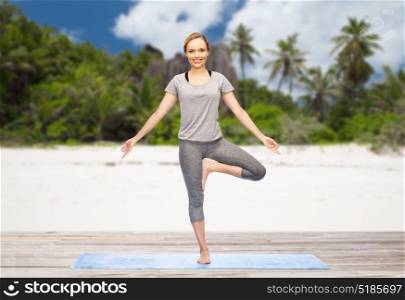 fitness, sport and healthy lifestyle concept - woman doing yoga in tree pose on mat over exotic tropical beach background. woman doing yoga in tree pose on beach