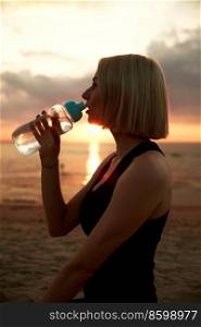 fitness, sport, and healthy lifestyle concept - woman drinking water from bottle on beach over sunset. woman drinking water from bottle on beach