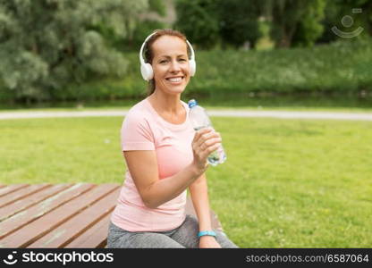 fitness, sport and healthy lifestyle concept - woman in headphones drinking water after exercising in park. woman in headphones drinking water in park