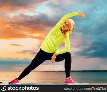 fitness, sport and healthy lifestyle concept - woman stretching on exercise mat over sea sunset background. woman stretching on exercise mat at seaside