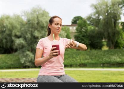 fitness, sport and healthy lifestyle concept - woman with smoothie or shake in plastic cup looking at smart watch in park. woman with shake looking at smart watch in park