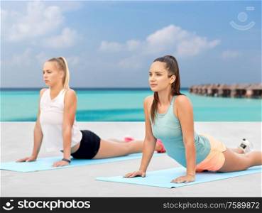 fitness, sport, and healthy lifestyle concept - women training or doing yoga on exercise mats over sea and bungalow on background. women doing sports on exercise mats outdoors